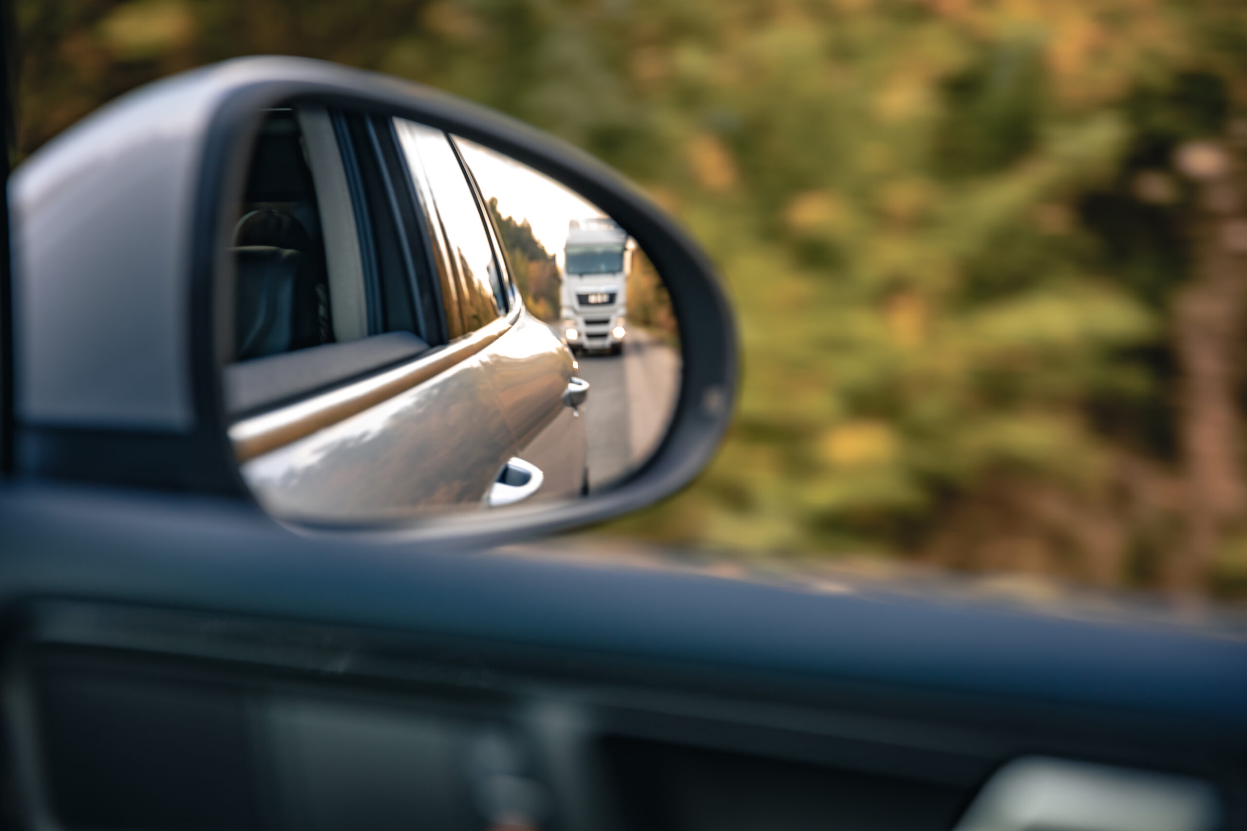 Vue dans un rétroviseur latéral montrant un camion qui suit à distance sur une route bordée d'arbres, illustrant l'importance de surveiller les véhicules derrière soi pour maintenir les distances de sécurité.
