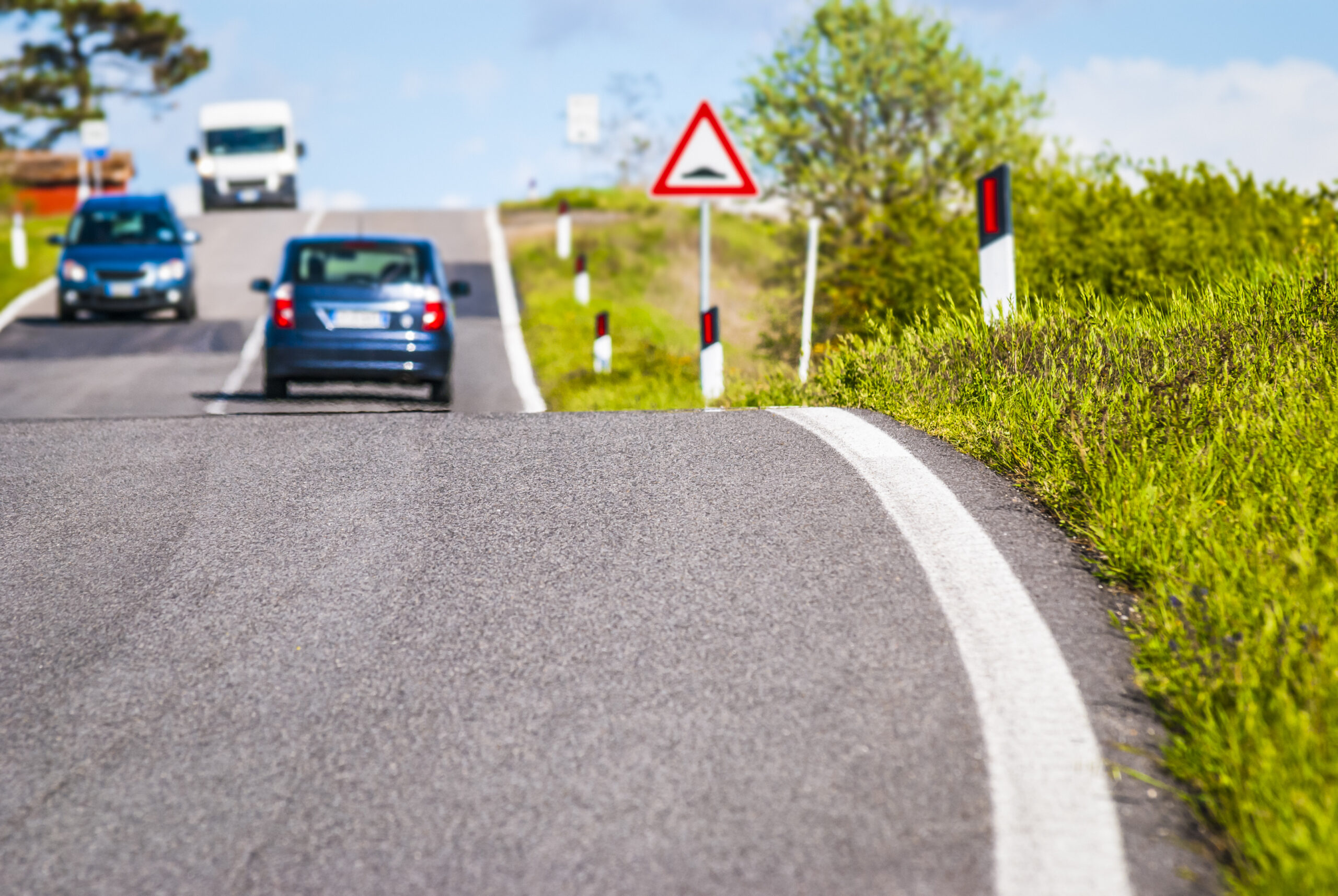 Plusieurs voitures circulent sur une route de campagne légèrement vallonnée, avec des panneaux de signalisation visibles et des marquages au sol, illustrant l’importance du respect des distances de sécurité en conditions de circulation normales.