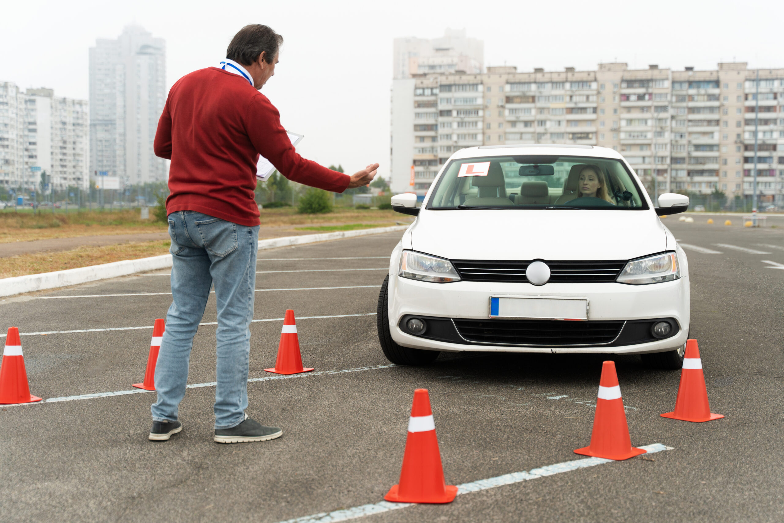 Instructeur de conduite en pull rouge guidant une conductrice dans une Volkswagen blanche sur un parcours d'entraînement délimité par des cônes orange, démontrant l'apprentissage pratique des distances de sécurité et du contrôle du véhicule.