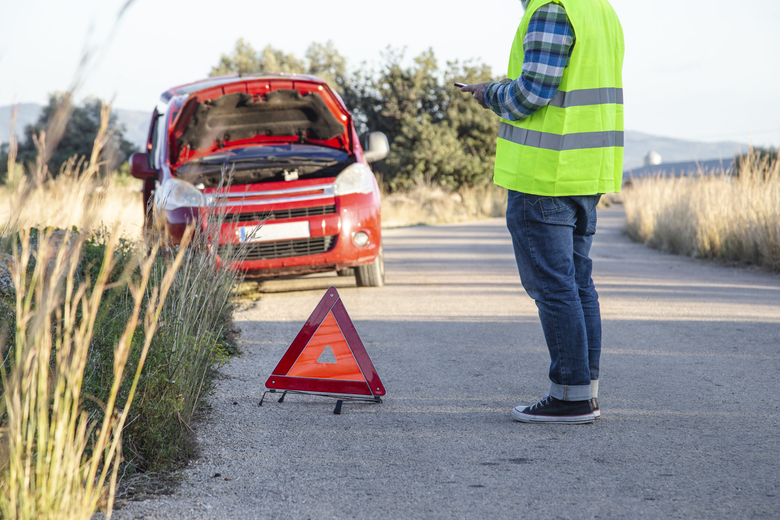 Conducteur en gilet jaune de sécurité près d’une voiture en panne avec capot ouvert, triangle de signalisation placé sur la chaussée – illustration des équipements obligatoires en voiture en 2025.