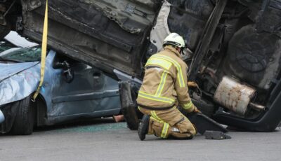 Pompier en tenue de protection agenouillé près d'une voiture accidentée retournée sur un autre véhicule.