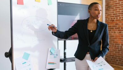 Femme en tailleur noir présentant un schéma sur un tableau blanc avec le mot Budget au centre dans un bureau.